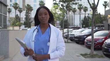 Genç Afrikalı Amerikalı kadın doktorun elinde dizüstü bilgisayarla hastanenin önünde durup gülümsediğini gösteren video.