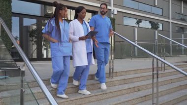 Doktor ve mavi önlüklü hemşirelerin hastane binasının merdivenlerinden tabletle inerken videosu.