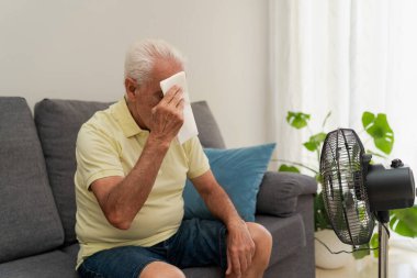 Evdeki koltukta elektrikli vantilatörle otururken alnından ter silen yaşlı bir adam.