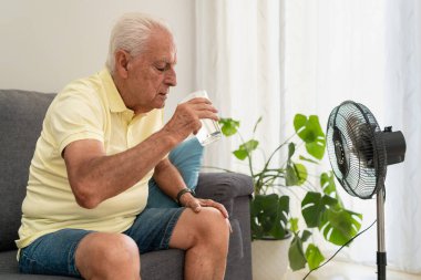 Kıdemli adam sıcak havada evde kanepede otururken vantilatörle su içip serinliyor.