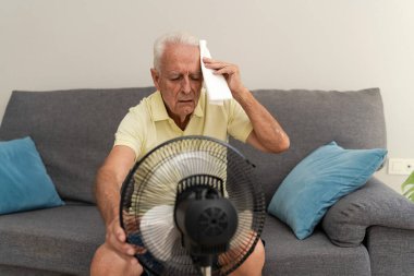 Yaşlı adam elektrikli yelpazeyle serinliyor ve alnındaki teri siliyor yaz sıcağından acı çekiyor.