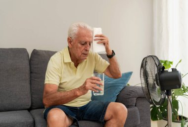 Evdeki elektrikli vantilatörün önünde otururken terleyen ve cam su tutan son sınıf öğrencisi.