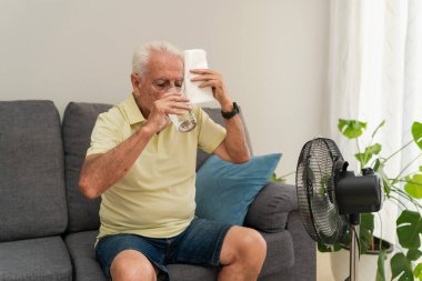 Son sınıf öğrencisi içme suyu içiyor ve elektrikli vantilatörle kanepede oturmuş terini havluyla siliyor.