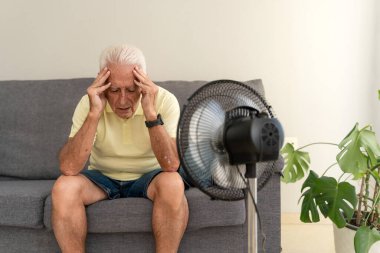 Yorgun düşmüş yaşlı bir adam evde yaz sıcağında elektrikli vantilatörle serinlemeye çalışıyor.