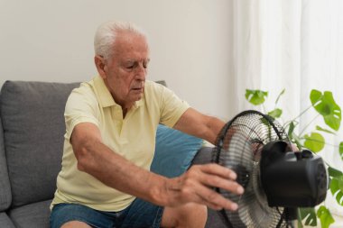 Son sınıf öğrencisi sıcak yaz günlerinde kanepede otururken evde elektrikli vantilatörle ferahlatıyor.