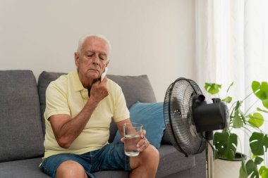 Son sınıf öğrencisi kanepede oturmuş evdeki vantilatörün yanında bir bardak su tutarak yüzünden ter siliyor.