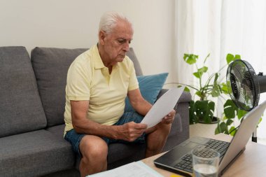 Evdeki kanepede dizüstü bilgisayar ve belgeler kullanarak ev işleriyle ilgilenen kıdemli bir adam.