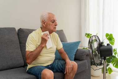 Son sınıf öğrencisi kanepede oturmuş terliyor, evinde elektrikli vantilatör kullanarak kendini sıcak hissediyor.