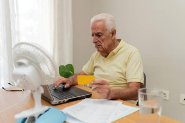 Çevrimiçi ödeme için dizüstü bilgisayar ve kredi kartı kullanan yaşlı bir adam, yaz sıcağı sırasında vantilatörden gelen serinletici hava akımının keyfini çıkarıyor