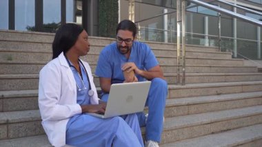 Hastanenin merdivenlerinde dizüstü bilgisayar kullanarak oturan iki farklı sağlık çalışanının videosu.