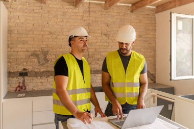 İki inşaat işçisi ev tadilatı sırasında planlar ve dizüstü bilgisayarlar hakkında konuşuyor.