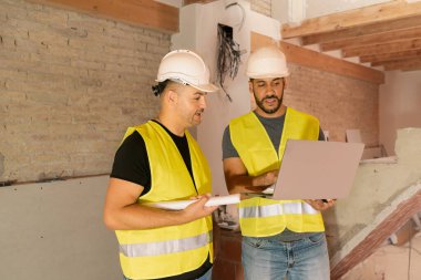 Ev tadilatı sırasında dizüstü bilgisayar ve plan kullanan iki erkek inşaat işçisi.