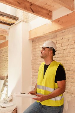 Güvenlik yeleği ve kask giyen erkek inşaat işçisi yenileme sahasında ahşap tavanı denetliyor.