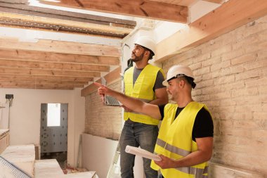 İki erkek inşaat işçisi dizüstü bilgisayar ve planlarla yenileme alanındaki tavan yapısını inceliyor.