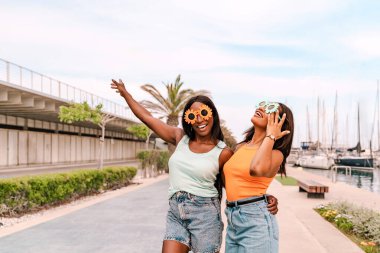 Güneş gözlüğü takan iki neşeli çok ırklı kadın liman yakınlarında yaz tatilinin tadını çıkarıyor.
