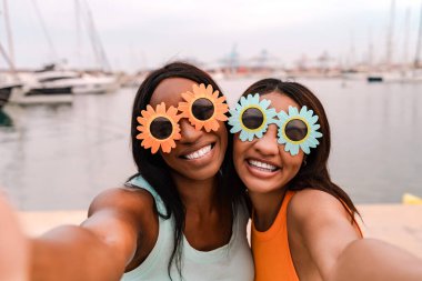 Çiçek şeklinde güneş gözlüğü takan ve deniz kenarında selfie çeken iki mutlu kadın arkadaş.