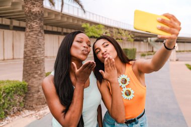 İki çok ırklı bayan arkadaş kameraya öpüşürken akıllı telefonuyla selfie çekiyorlar.