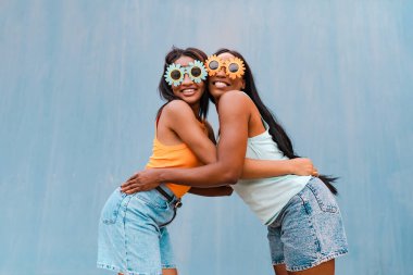 Komik güneş gözlüğü takmış, mavi arka planda birbirine sarılan iki çok kültürlü kadın.
