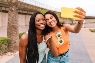 İki neşeli çoklu etnik kadın güneşli bir günde akıllı bir telefonla selfie çekerken eğleniyor.