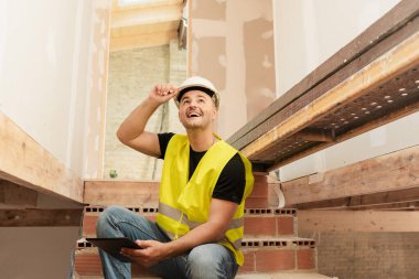 Güler yüzlü inşaat işçisi, güvenlik yeleği ve inşaat alanındaki merdivenlerde oturuyor.