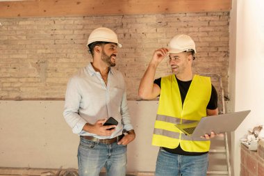 Güler yüzlü inşaat işçisi ve mimar güvenlik başlığı ve yelek içinde dizüstü bilgisayar kullanarak projeyi tartışıyorlar