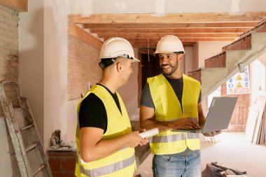 Ev tadilatında çalışan iki güvenlik yelekli ve kasklı inşaat işçisi dizüstü bilgisayar kullanıyor.