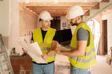 İnşaat alanında dizüstü bilgisayarla bina planlarını analiz eden iki inşaat işçisi.