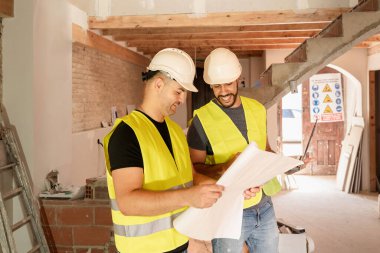 İki inşaat işçisi güvenlik yelekleri ve baret takıyor. Ev tadilatı sırasında planı kontrol ediyorlar.
