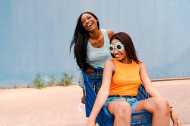 İki arkadaş gülüyor ve alışveriş arabasıyla oynuyorlar, komik gözlükler takıyorlar, açık havada yaz gününün tadını çıkarıyorlar.