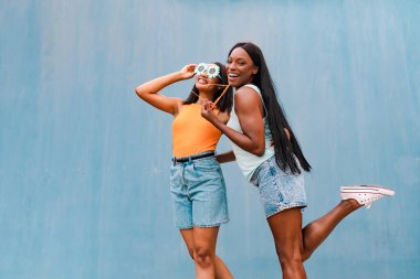 İki çok ırklı kadın komik güneş gözlüğü takıp, mavi arka planda gülümsüyor.