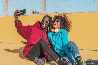 Neşeli çok ırklı arkadaşlar, parlak günlük kıyafetler ve güneş gözlüğü takıp, gülümsüyor ve güneşli bir günde spor sahasında takılırken akıllı telefondan selfie çekiyorlar.