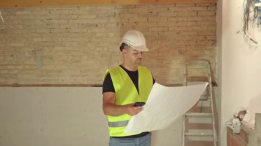 İnşaat alanının planlarına bakan sert şapkalı ve yansıtıcı yelekli erkek inşaat işçisinin videosu.