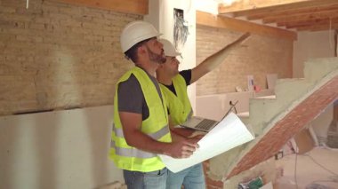 Sert şapkalı ve yelekli iki erkek inşaat işçisinin görüntüleri planları inceliyor ve bir inşaat alanında dizüstü bilgisayar kullanıyor.