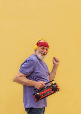 Sarı arka planda müzik kutusu ve kulaklıkla dans edip müzik dinleyen mutlu kıdemli adam.