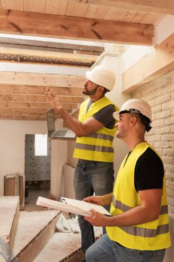 Ev inşaatında çalışan kasklı ve dizüstü bilgisayarı olan erkek inşaat işçileri.