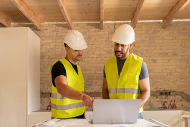 Ev yenileme projesi sırasında dizüstü bilgisayar kullanan güvenlik yeleği ve kask giyen iki inşaat işçisi