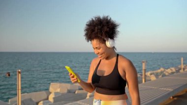 Virajlı İspanyol kadının spor giyim kuşağındaki videosu. Müzik dinliyor ve rıhtımda yürürken akıllı telefon kullanıyor.