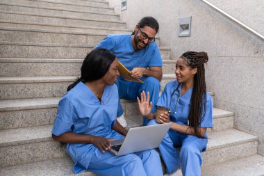 Çok ırklı tıp öğrencileri hastane merdivenlerinde dizüstü bilgisayar kullanarak birlikte çalışıyorlar.