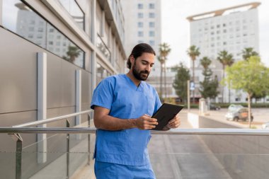 Mavi önlüklü erkek doktor hastanenin dışında dijital tablet, modern sağlık ve teknoloji kullanıyor.