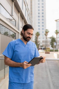 Mavi önlüklü genç erkek doktor hastane binasının dışında tablet bilgisayar kullanıyor.