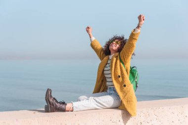 Sarı ceketli ve sırt çantalı genç kadın deniz kenarında kollarını kaldırıyor ve başarısını kutluyor.