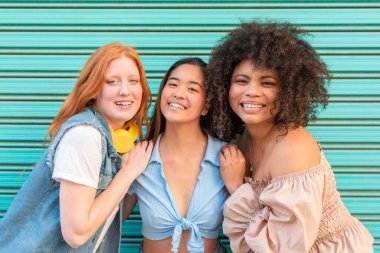 Kameraya gülümseyen turkuaz bir duvara sarılan üç neşeli çoklu etnik bayan arkadaş.