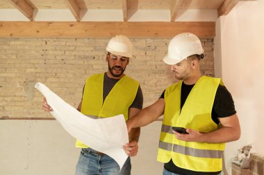 İki erkek inşaat işçisi yenileme alanının planlarını inceliyor.