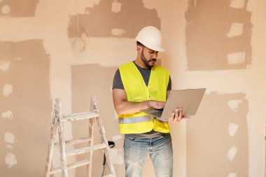 İnşaat sırasında güvenlik yeleği ve kask giyen erkek mimar dizüstü bilgisayar kullanıyor.