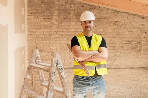 Erkek inşaat işçisi çelik yelek giyiyor ve kasketli. Seviye aracı ile poz veriyor ve inşaat alanında çapraz kollar kullanıyor.