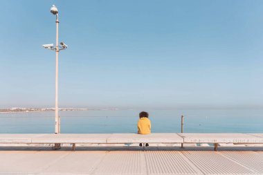 Sarı montlu bir kadının, bir bankta oturmuş, şehir denizini düşünürken dikiz görüntüsü.