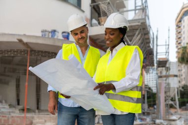 İki farklı güvenlik yeleği ve baret mühendisi inşaat projesini tartışan inşaat alanının planlarını okuyorlar.