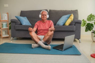 Yaşlı bir adam yoga minderinde kulaklık ve dizüstü bilgisayarla meditasyon yapıyor.