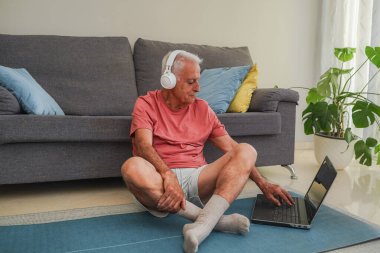 Evde yoga minderinde egzersiz yapan kulaklıklı son sınıf öğrencisi dizüstü bilgisayarla yaptığı antrenmanı takip ediyor.