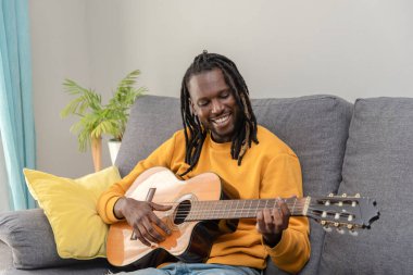 Rastalı Afrikalı Amerikalı adam evdeki oturma odasında kanepede oturup gitar çalmayı öğreniyor.
